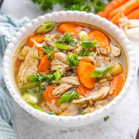 A close up shot of a bowl of chicken rice soup with big chunks of carrots and fresh green onions on top.
