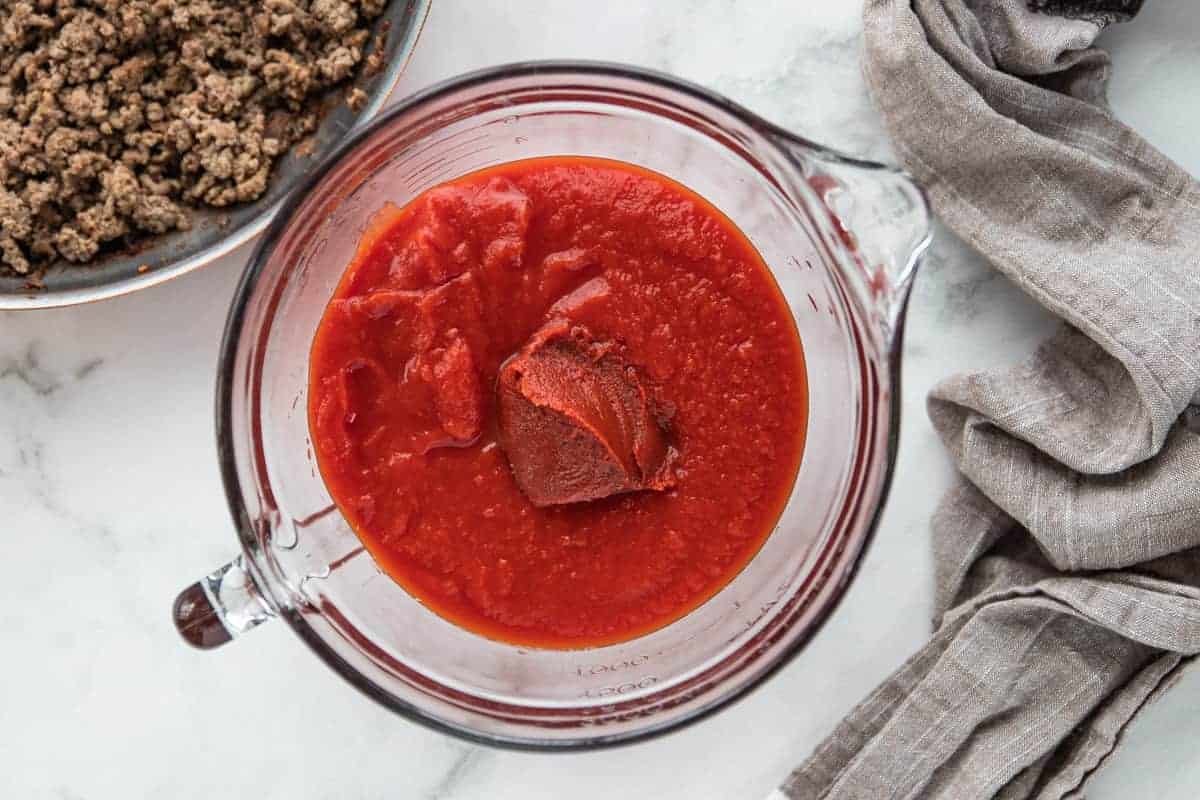 tomato sauce in a glass mixing bowl with browned meat in a skillet off to the side all on top of a granite countertop.