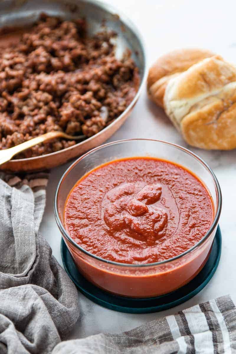 Sloppy Joe Mix showing sauce in a glass container, browned meat in a skillet and a toasted bun all on top of a granite countertop.