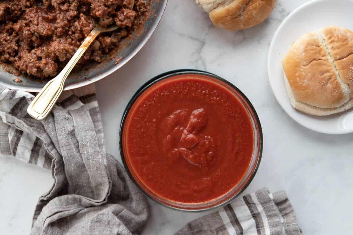 sauce, browned meat in a skillet and a bun on a plate all on top of a granite countertop.