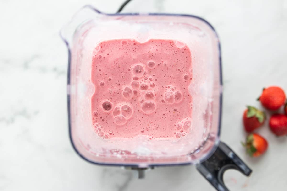 strawberry smoothie in the blender with strawberries on the side all on top of a granite countertop.
