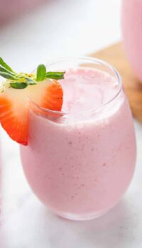 A small clear glass on a white counter filled with strawberry smoothie with a slice of strawberry on top.