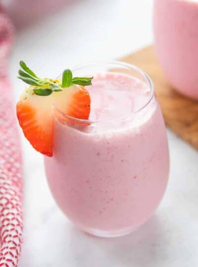 A small clear glass on a white counter filled with strawberry smoothie with a slice of strawberry on top.