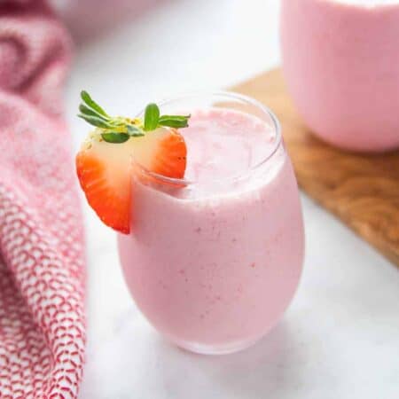 A small clear glass on a white counter filled with strawberry smoothie with a slice of strawberry on top.