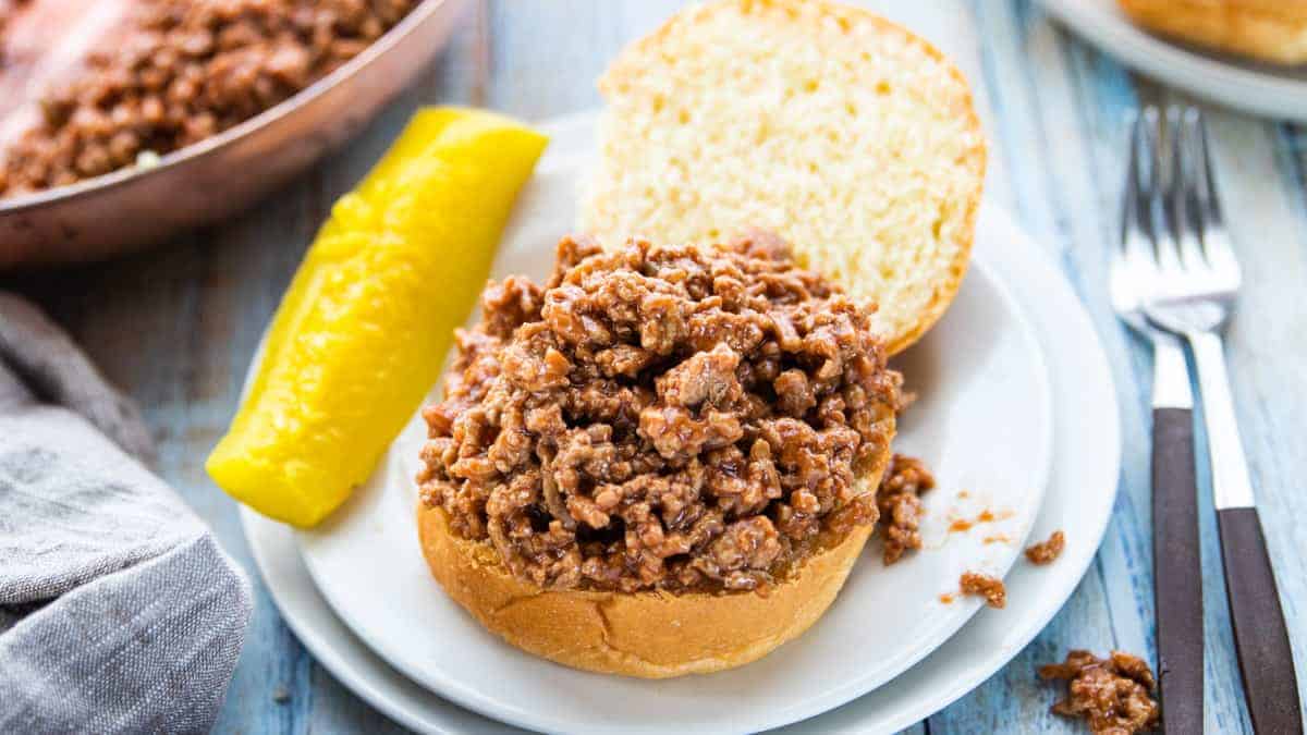 an open faced sloppy Joe sandwich on a white plate with a pickle on the side and a fork next to the plate all on top of a wooden countertop.