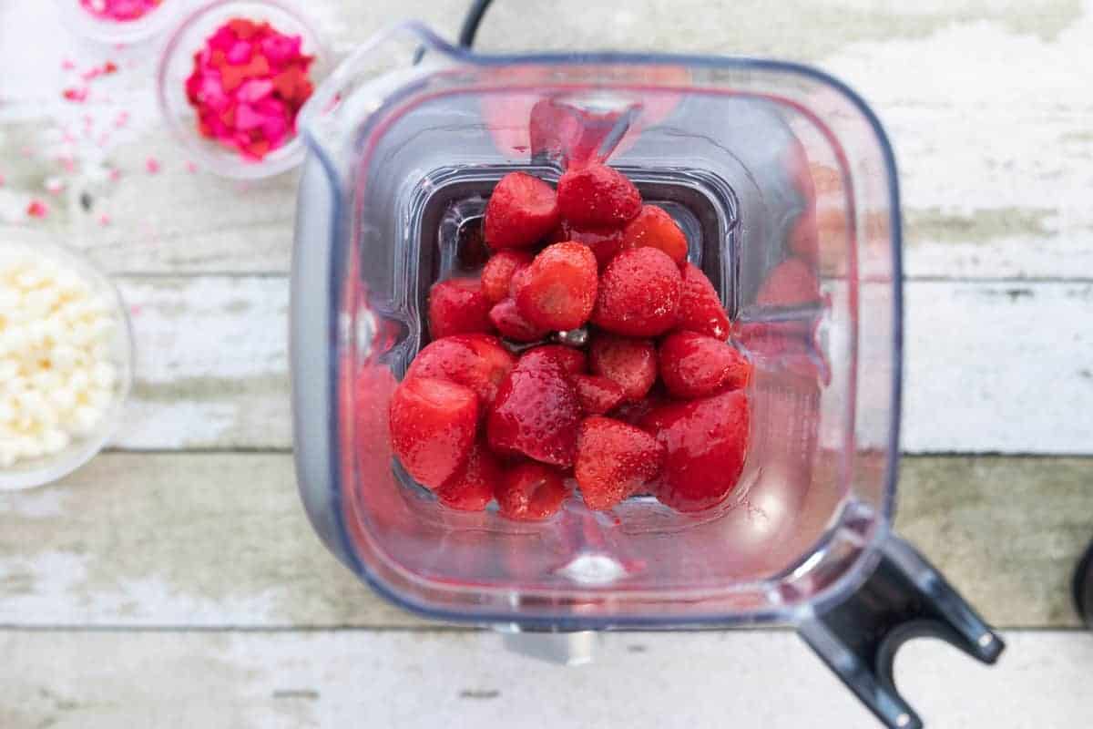 strawberries in a blender with other ingredients in small class bowls all on top of a wooden countertop.