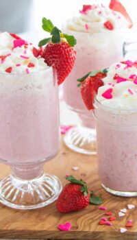 3 clear glasses filled with pink hot chocolate with strawberries, whipped cream, and sprinkles on top.