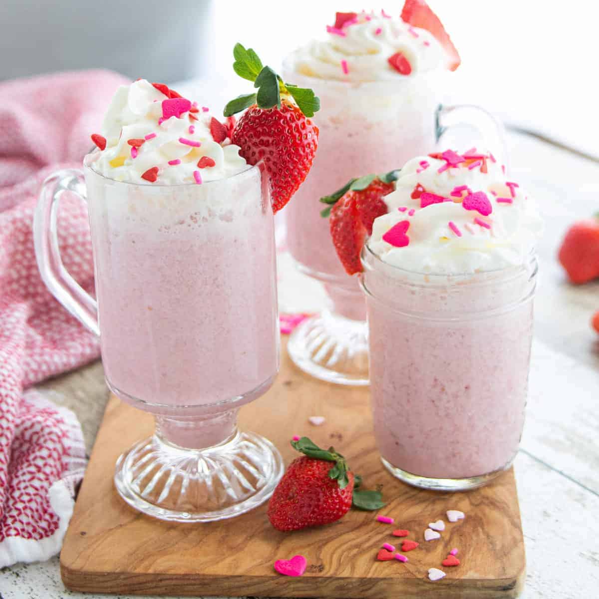 three glass mugs on top of a wooden board filled with pink hot chocolate topped with whipped cream.