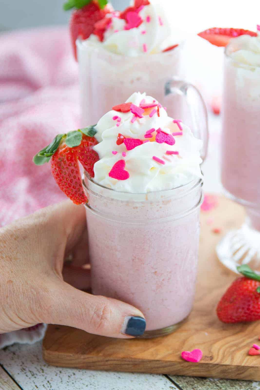 a hand holding a glass of pink hot chocolate with whipped cream and sprinkles with a strawberry.