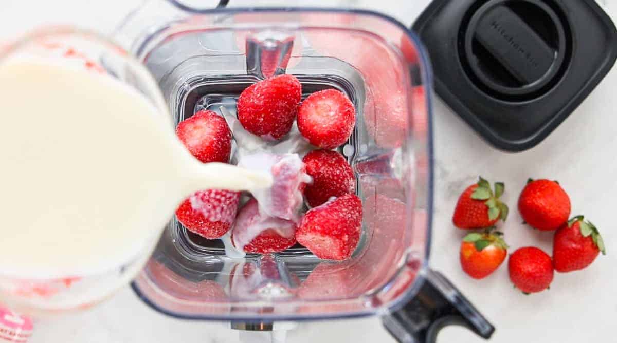 strawberries added to the blender with milk being poured into the blender.