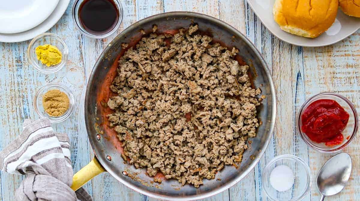 a skillet with browned ground turkey with small glass bowls of ingredients surround the skillet all on top of a wooden countertop.