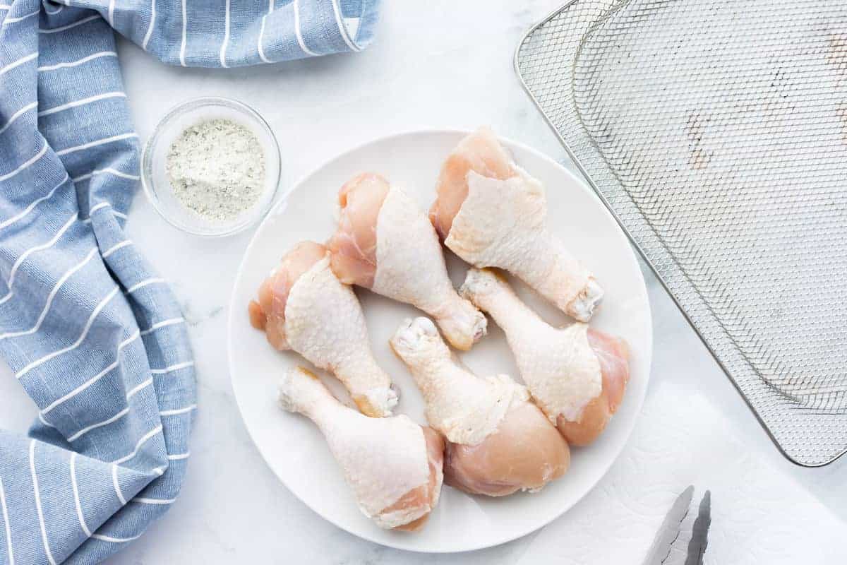 Air Fryer Drumsticks showing raw chicken legs on a white plate with seasoning in a white glass bowl.