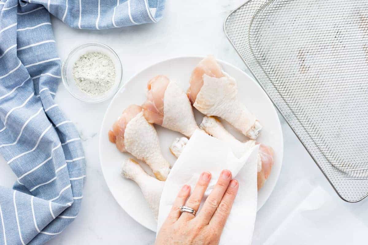 Air Fryer Chicken Legs showing raw chicken legs on a serving platter with a hand holding a paper towel and patting each chicken drumstick dry.