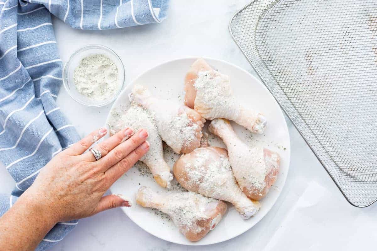 Air Fryer Drumsticks showing raw chicken legs on a white plate with seasoning in a white glass bowl.