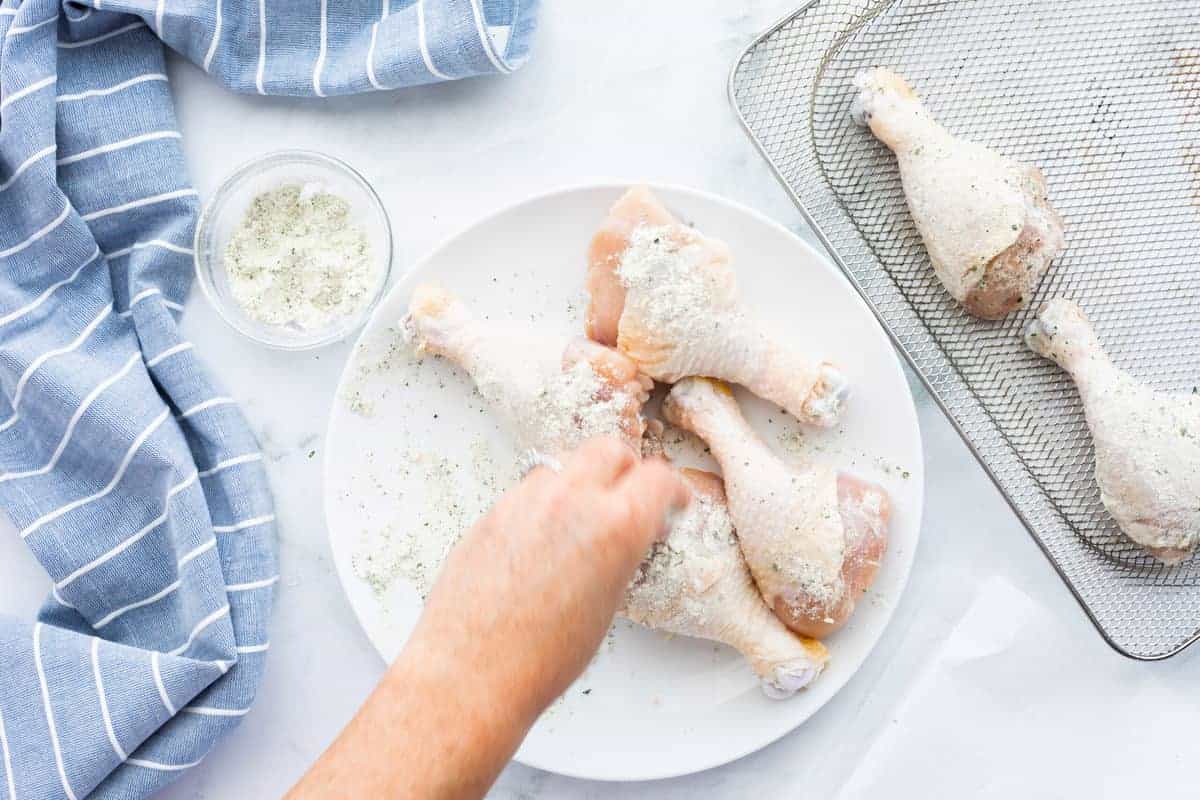 Air Fryer Chicken Legs showing a hand sprinkling seasoning over raw chicken legs with a few seasoning chicken drumsticks on the air fryer tray.