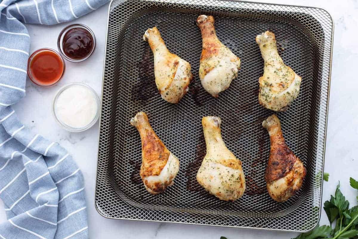 Air Fryer Chicken Legs showing air fried chicken drumsticks on an air fryer tray with multiple dipping sauces on the side of the tray.
