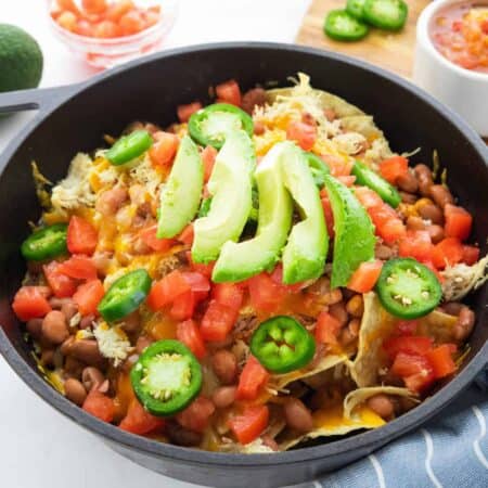 skillet nachos shown in a cast iron skillet with diced tomatoes, jalapeños, beans, and avocados on top.