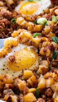 A close up of a cooked skillet scramble showing diced potatoes, bacon bits, cheese, and a fried egg on top.