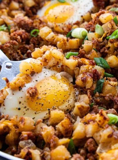 A close up of a cooked skillet scramble showing diced potatoes, bacon bits, cheese, and a fried egg on top.