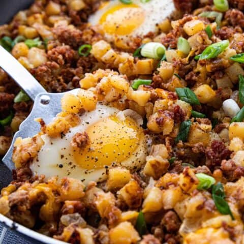A close up of a cooked skillet scramble showing diced potatoes, bacon bits, cheese, and a fried egg on top.
