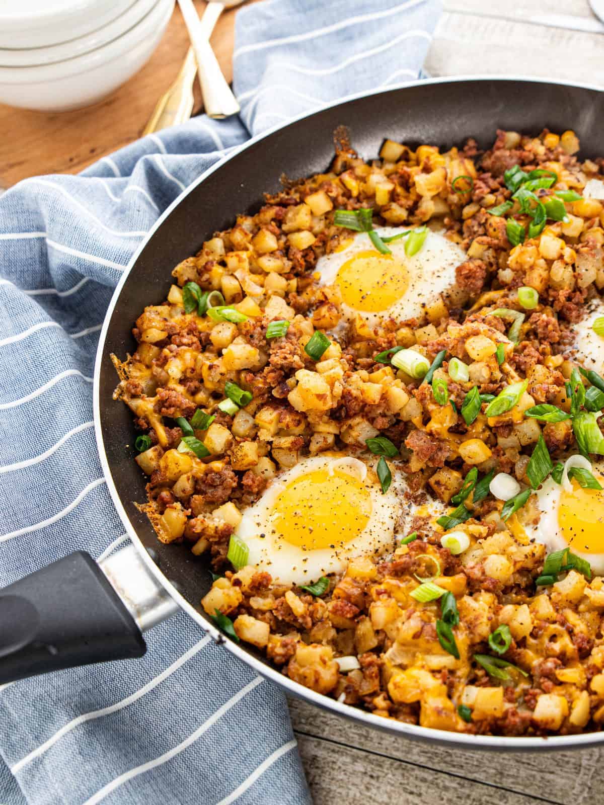 A sausage cowboy skillet is shown with 4 fried eggs, diced potatoes, and green onion garnish next to a blue stripped napkin. 