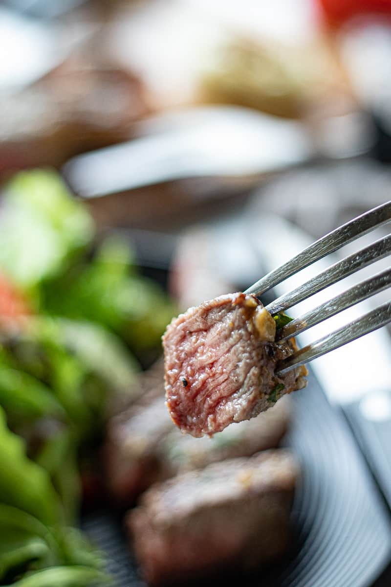 A close up of a bite s sliced steak on a fork with the rest of the plate blurry in the background.