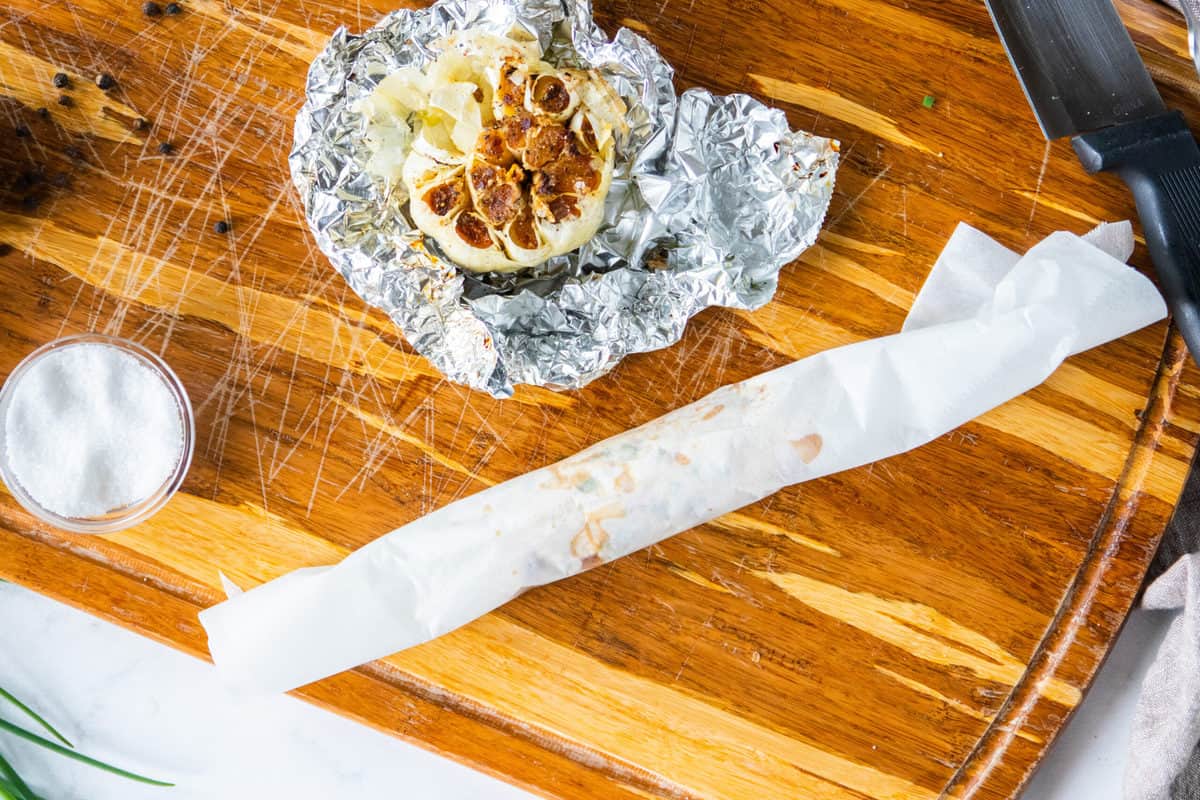 compound butter is wrapped in white wax paper on a cutting board with a head of roasted garlic.