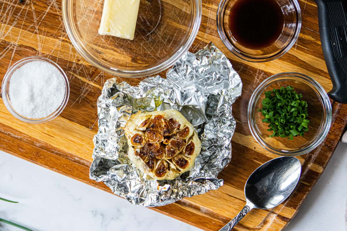 A top down view of a roasted head of garlic on tin foil pulled back next to butter, salt, chives and a spoon.