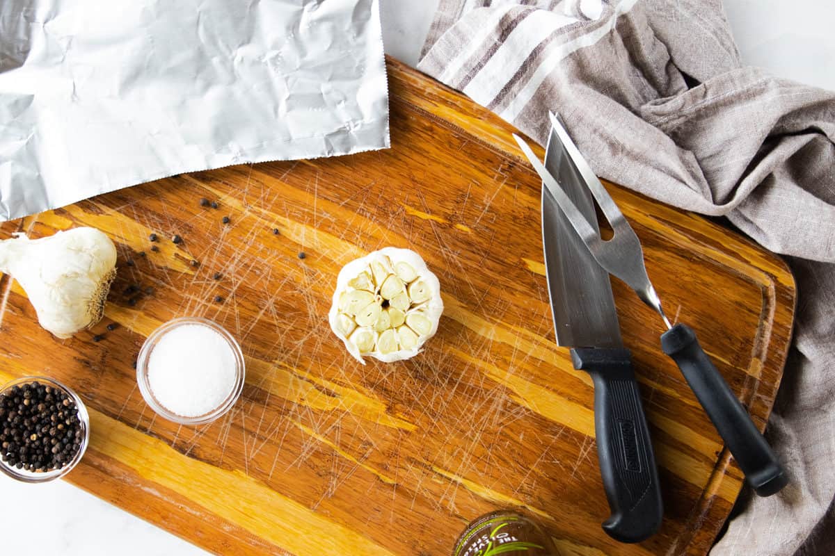 A top down view of a cutting board with a head of garlic with the top sliced off next to salt and pepper. 