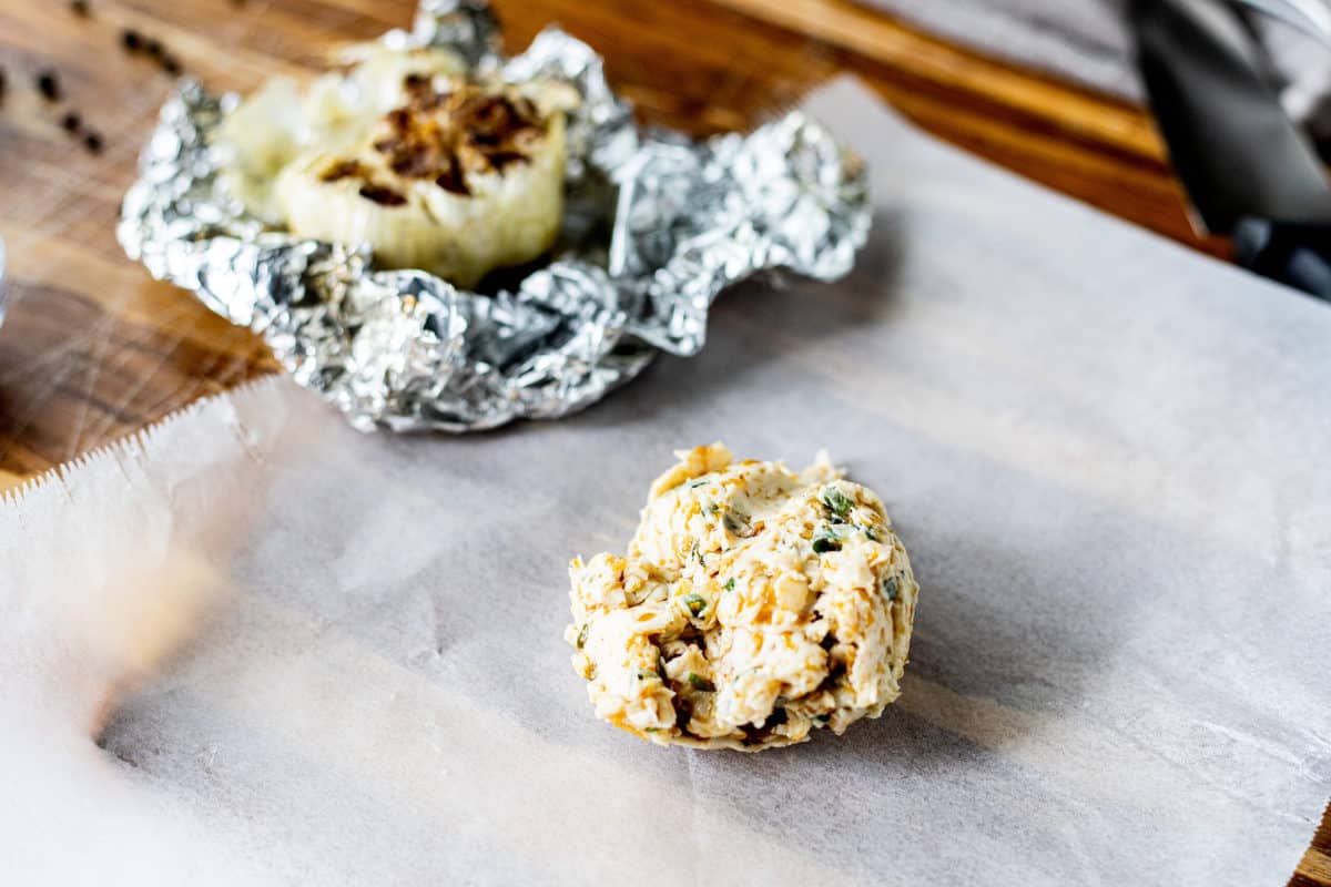 A ball of compound butter on a piece of wax paper with roasted garlic in the background.