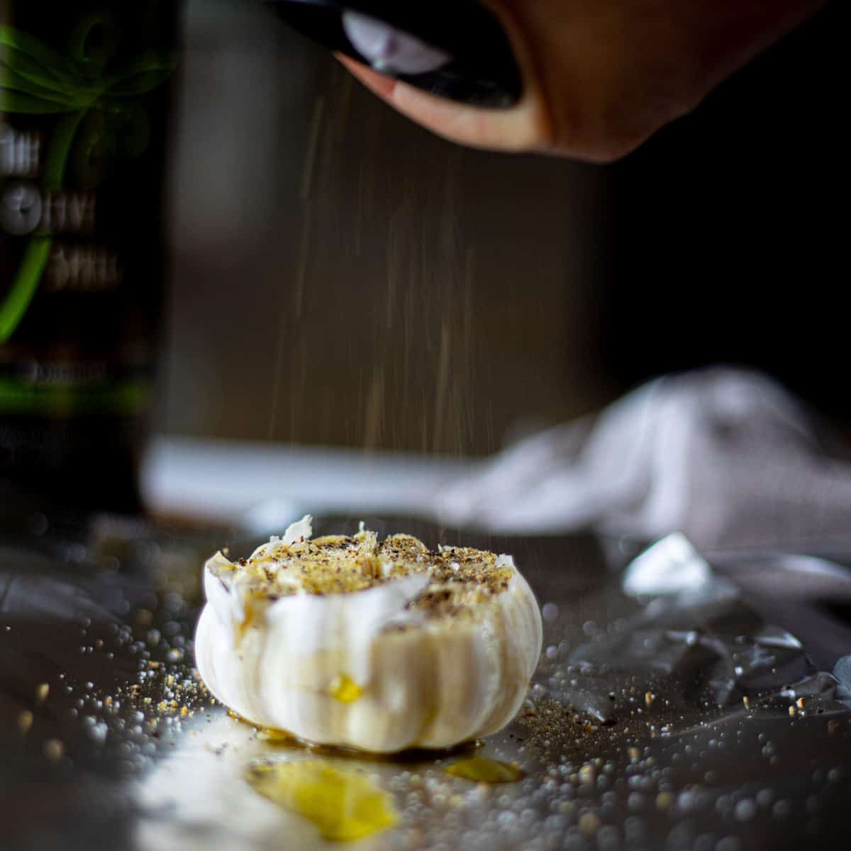 A head of garlic on tin foil with olive oil on it with a hand twisting cracked pepper onto it. 