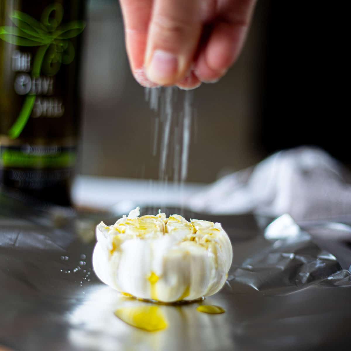 A head of garlic on tin foil with a hand sprinkling salt onto it. 