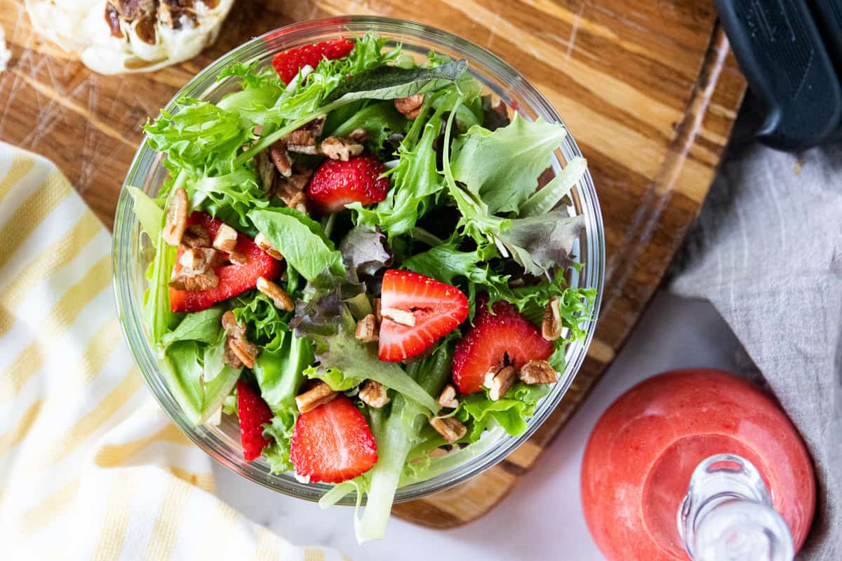 A top down view of a lettuce salad with sliced strawberries and pecans with strawberry vinaigrette in a glass jar next to it. 