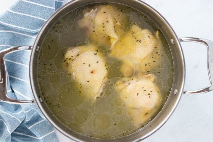 Boiled chicken thighs in a stainless steel stockpot with a blue and white towel next to the stockpot. 