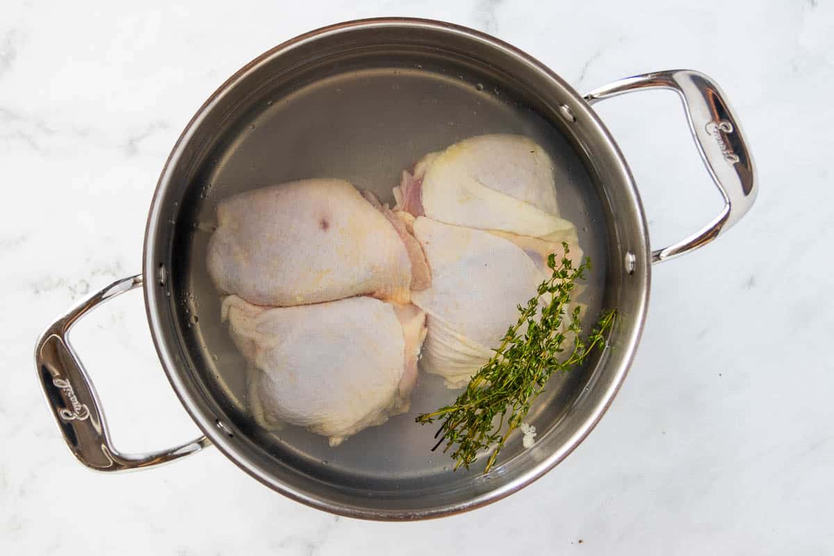 Raw chicken thighs in a stainless steel stockpot with salt and a sprig of thyme.
