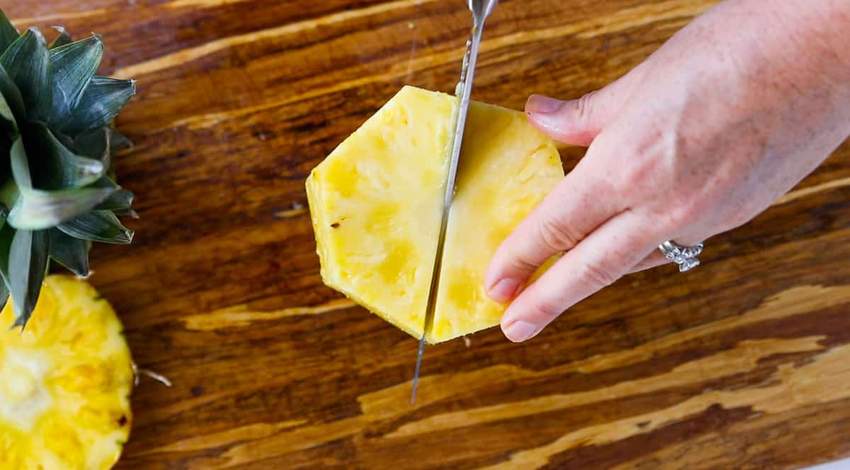 A peeled fresh pineapple on a cutting board is being cut in half.