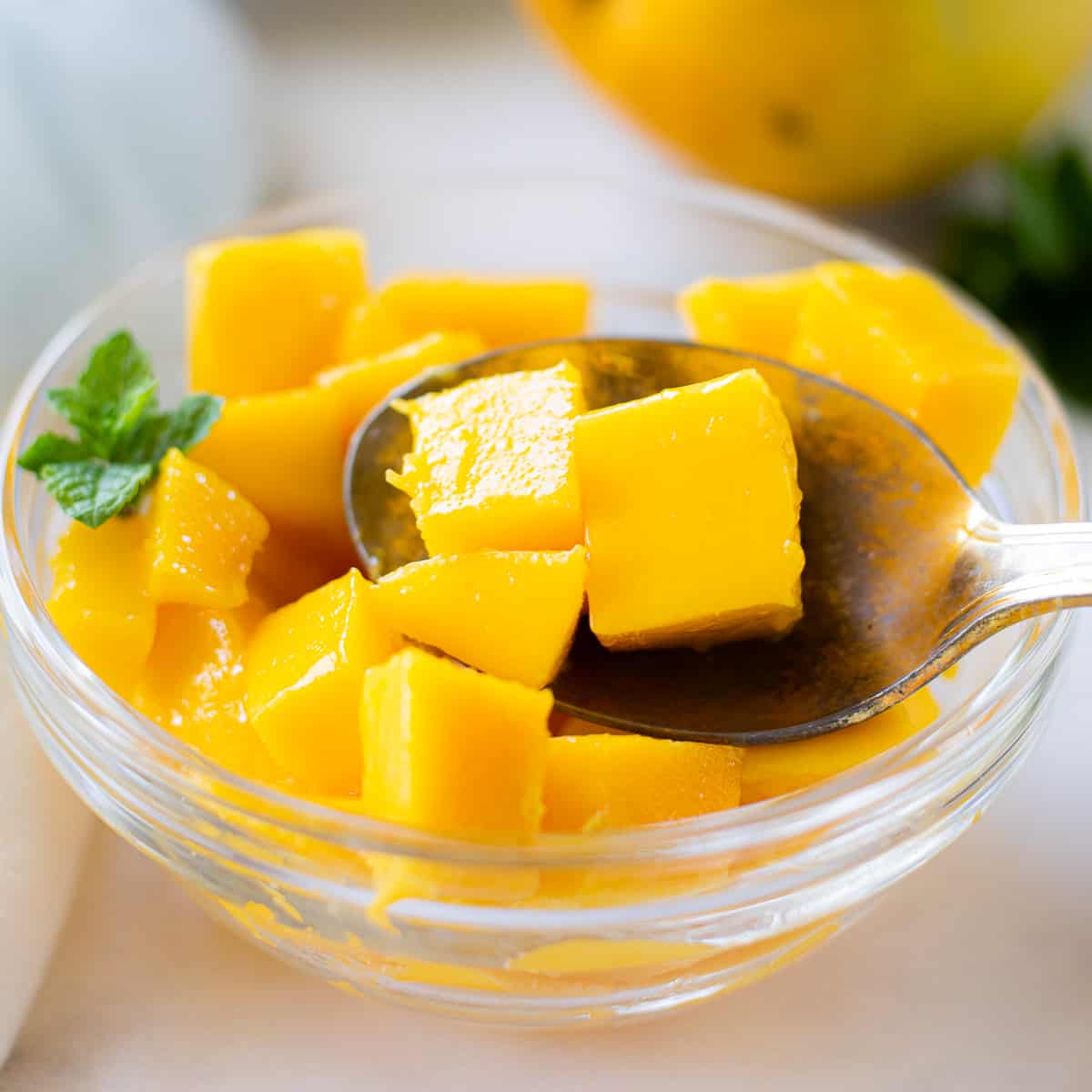 A clear bowl of diced mango chunks with a spoon holding 3 cubes with a mint garnish.