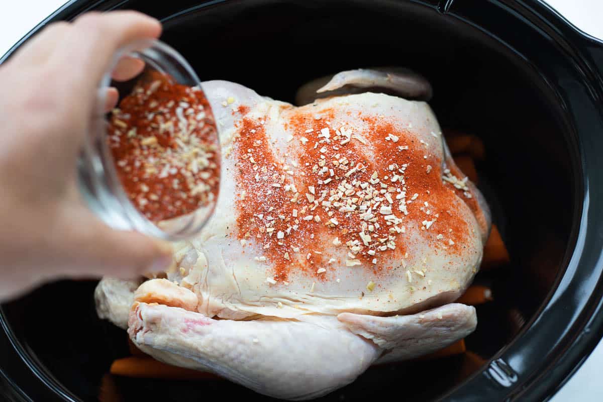 A hand pouring sprinkling a dry rub on top of a raw chicken that is placed on top of peeled carrots all in the base of a black slow cooker.
