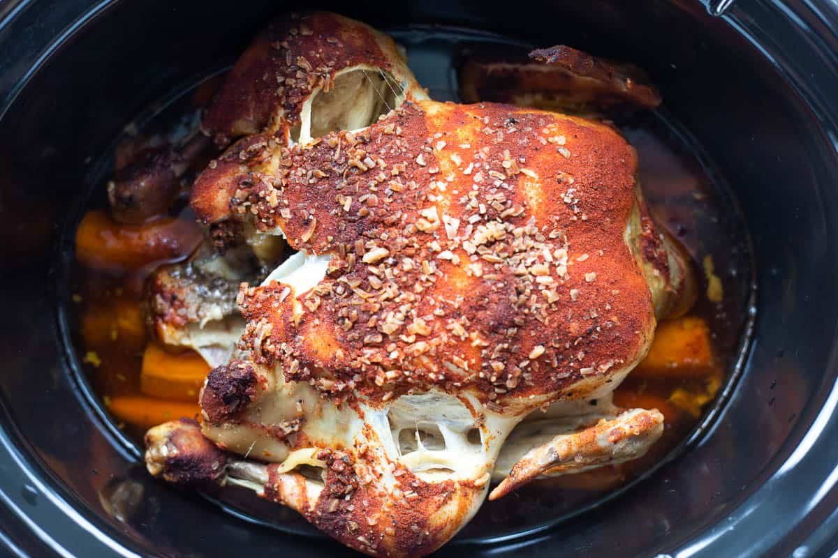A cooked whole chicken and cooked carrots in the base of a slow cooker. 