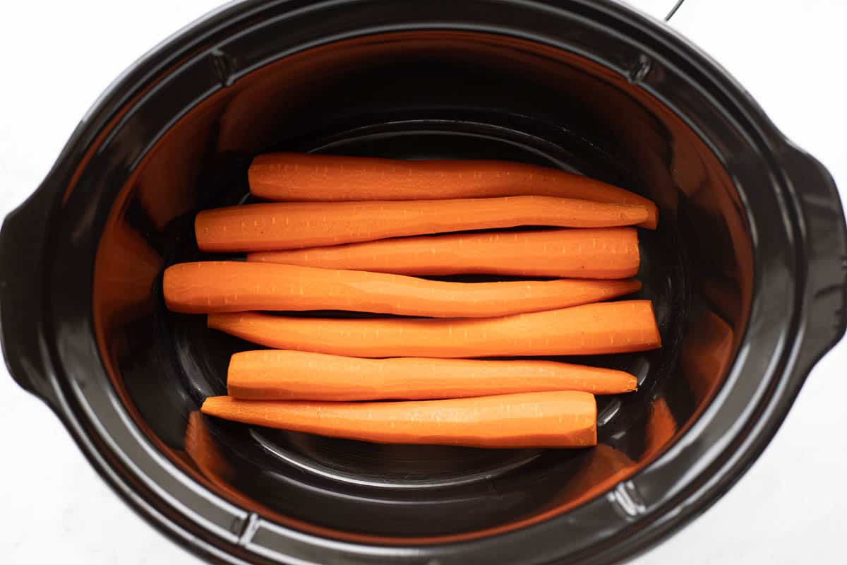 Peeled, fresh carrots placed in the base of a black slow cooker. 