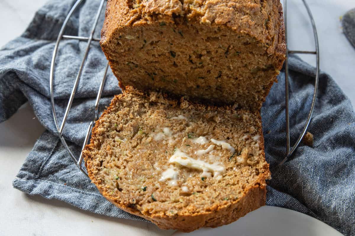 A piece of buttered banana zucchini bread sliced from the loaf all on top of a cooling rack that is on top of a blue dish towel. 