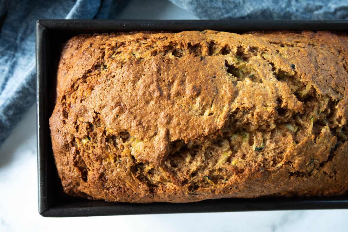 A loaf of banana zucchini bread  in a loaf pan on top a dish towel all on top of a granite countertop. 