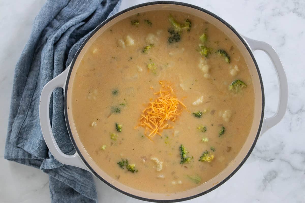 Broccoli cauliflower soup topped with cheddar cheese in a white stockpot on a granite countertop with a blue dish towel next to the pot.