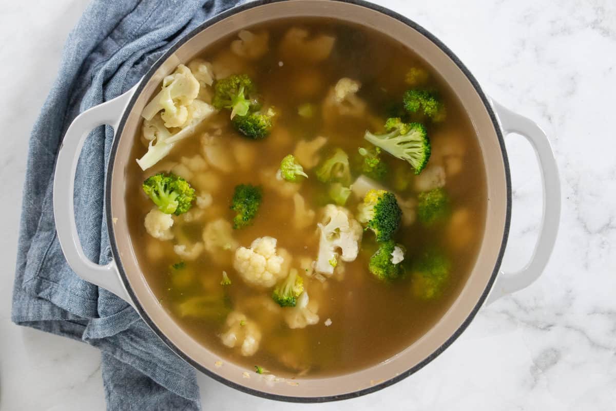 Broccoli and cauliflower florets covered with chicken broth in a white stockpot on a granite countertop with a blue dish towel next to the pot.