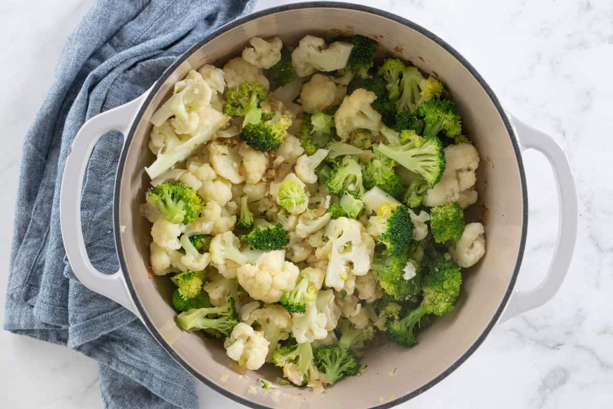 Broccoli and cauliflower florets mixed with chicken broth in a white stockpot on a granite countertop with a blue dish towel next to the pot.