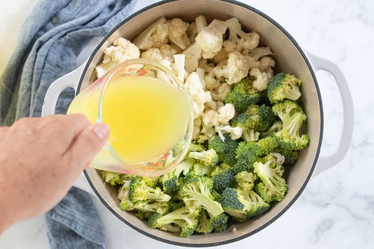 Broccoli and cauliflower florets in a white stockpot with chicken broth being poured from a measuring cup into the pot.