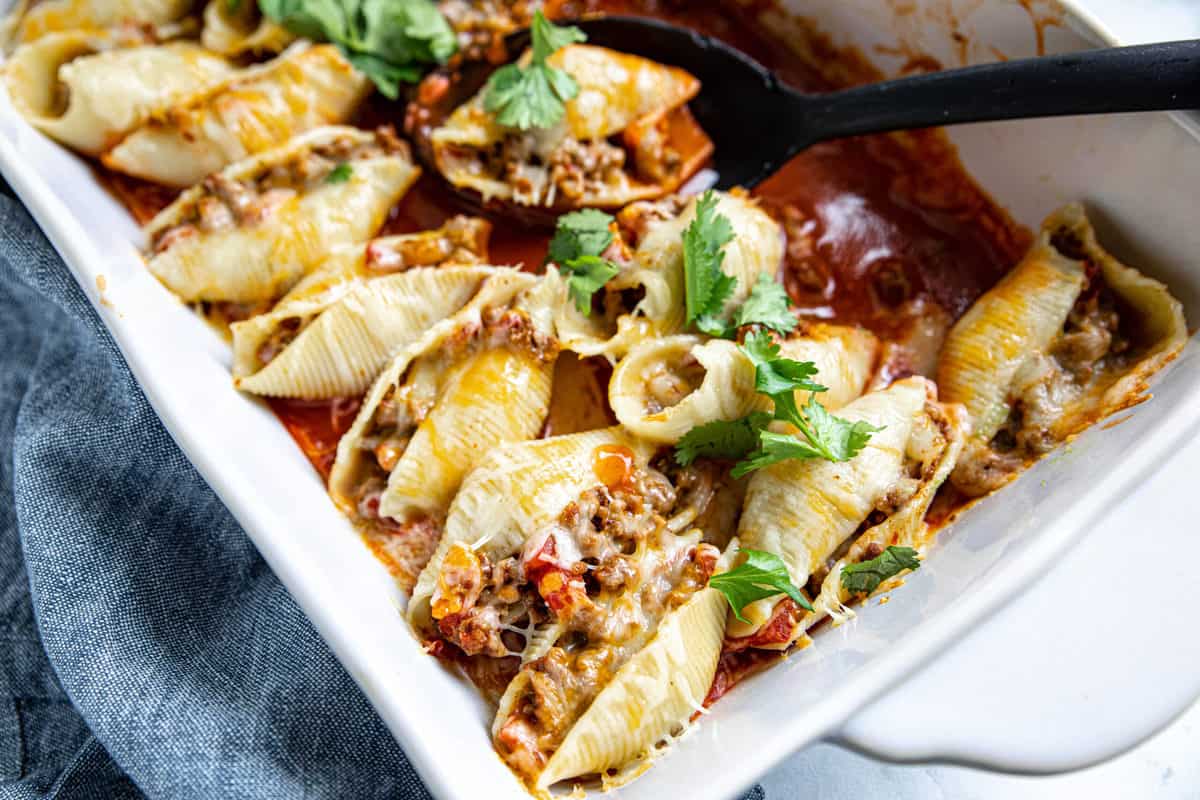Mexican Stuffed Shells baked in a white casserole dish with a spoon ladling out a shell. 