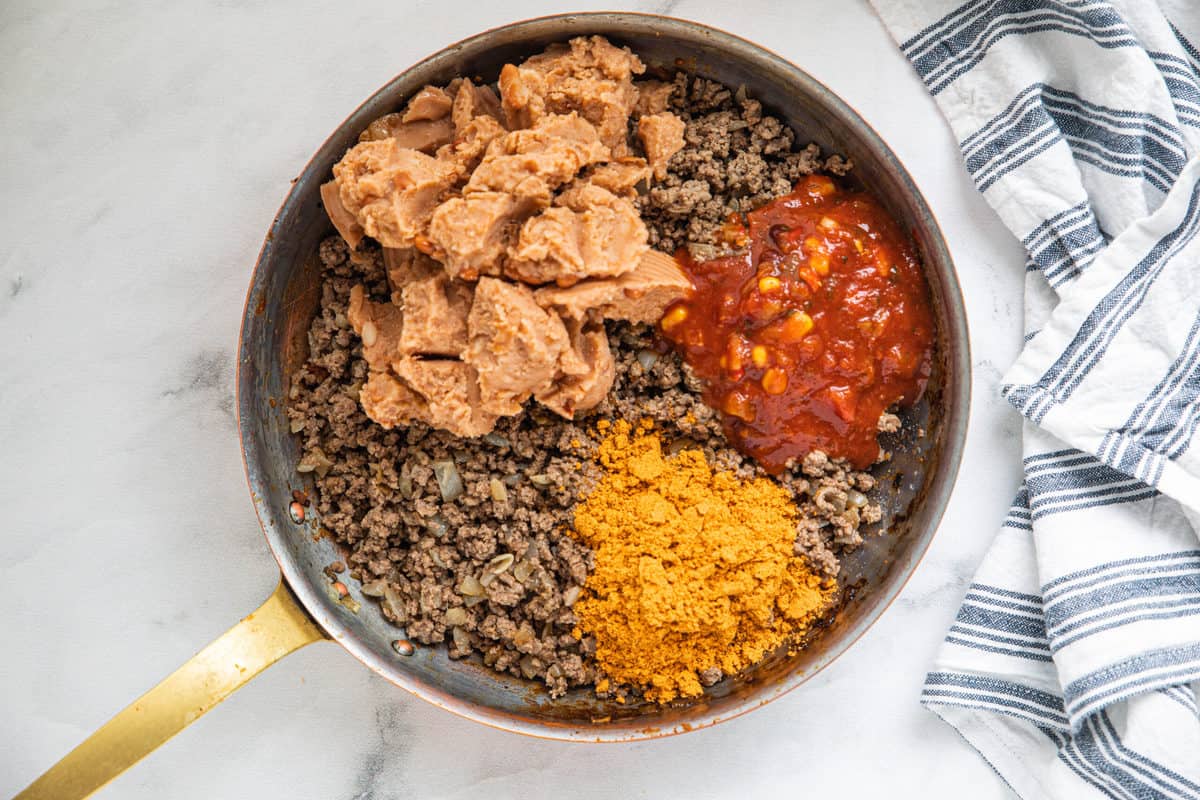 Ground beef, refried beans, salsa and taco seasoning being shown in a large skillet on a granite countertop with a dish towel next to the skillet.