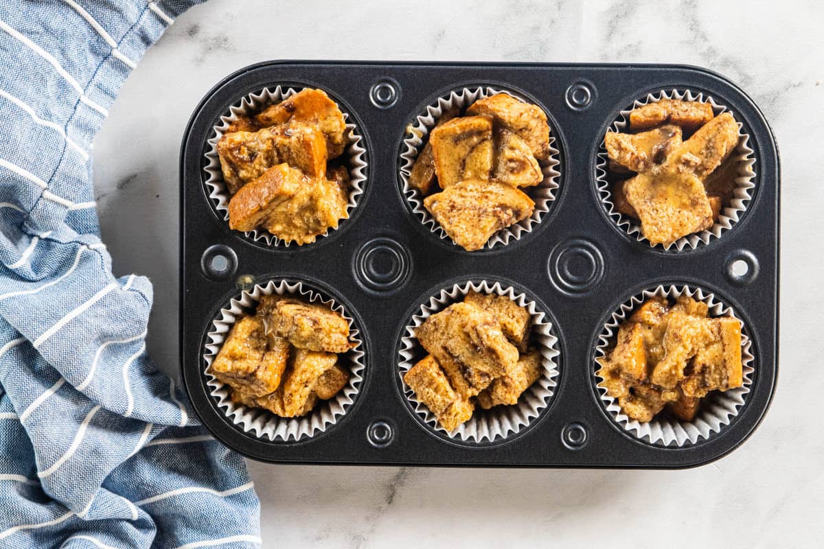 Baked French toast muffins in a muffin tin with a blue dish towel next to the tin all on top of a countertop.