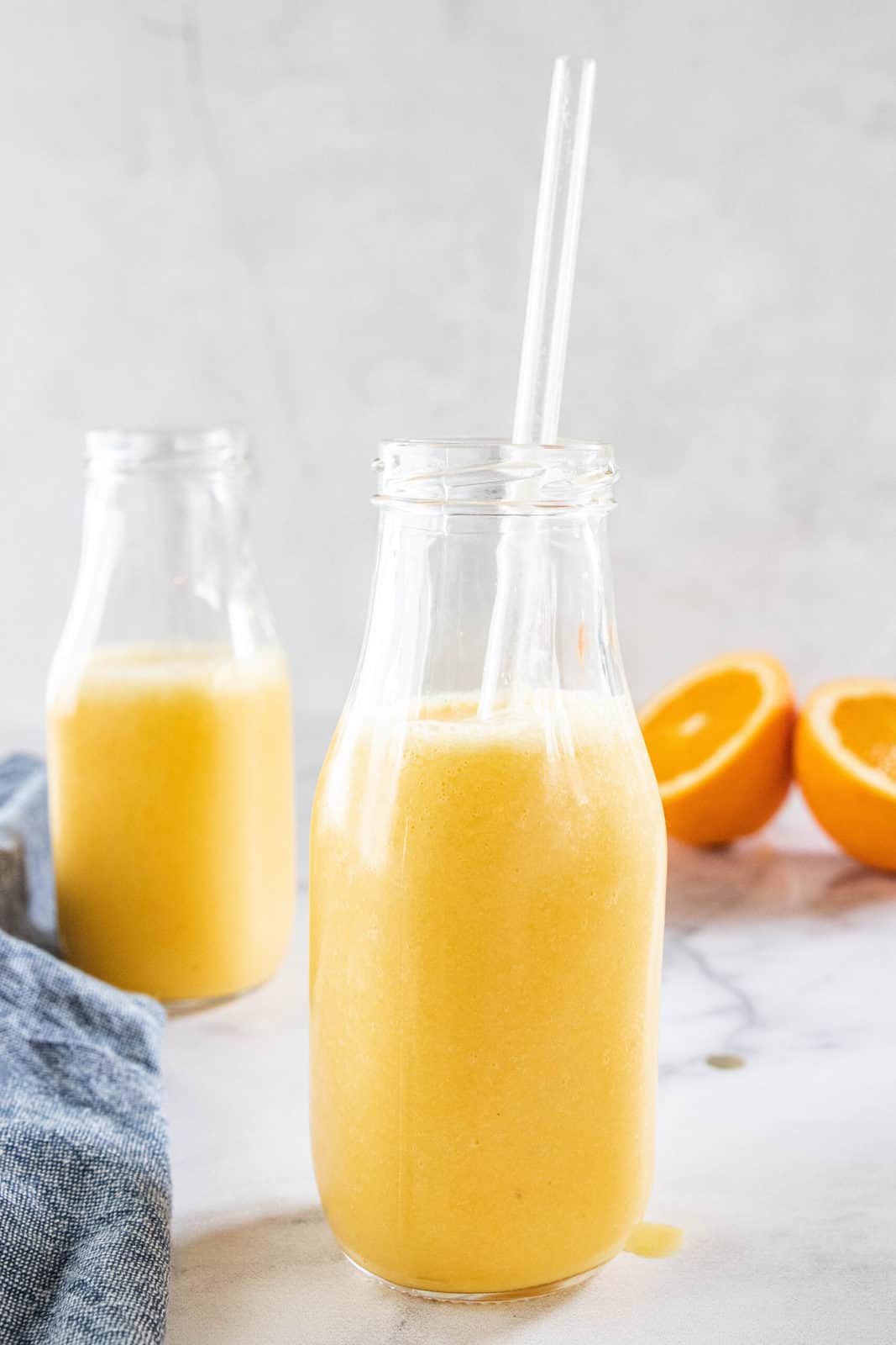 Two jars filled with an orange smoothie with orange halves and a dish towel next to the jars all on top of a granite countertop.